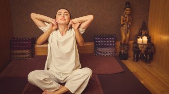 Thai massage stretch. Relaxed young woman is sitting on floor in lotus position. Masseuse is touching her arms